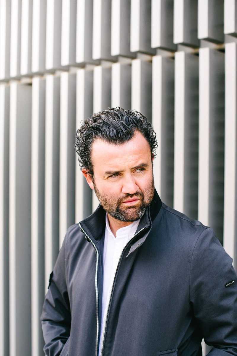 Photo of Daniel Mays in a black jacket looking into the distance with a textured building behind him.