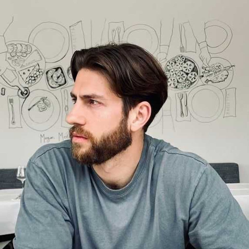 A white man with dark brown hair and a styled beard. Looking away from the camera. 