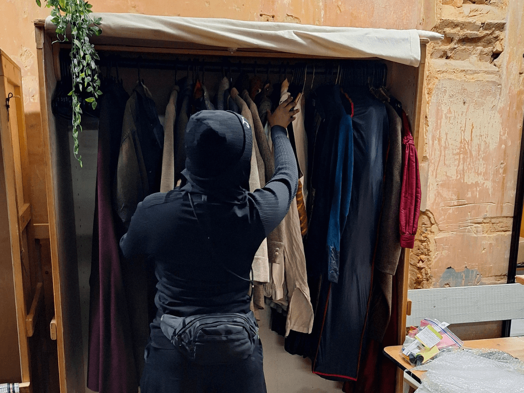Wardrobe intern, Aisha, a black young woman wearing blacks - looking through the wardrobe at Alexandra Palace.