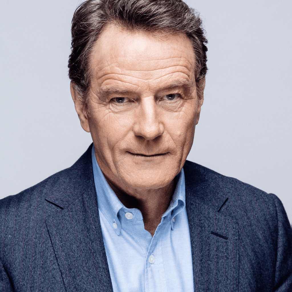 Headshot of Bryan Cranston, a man with neatly styled hair wearing a tailored dark blazer over a light blue shirt, set against a plain light grey background.