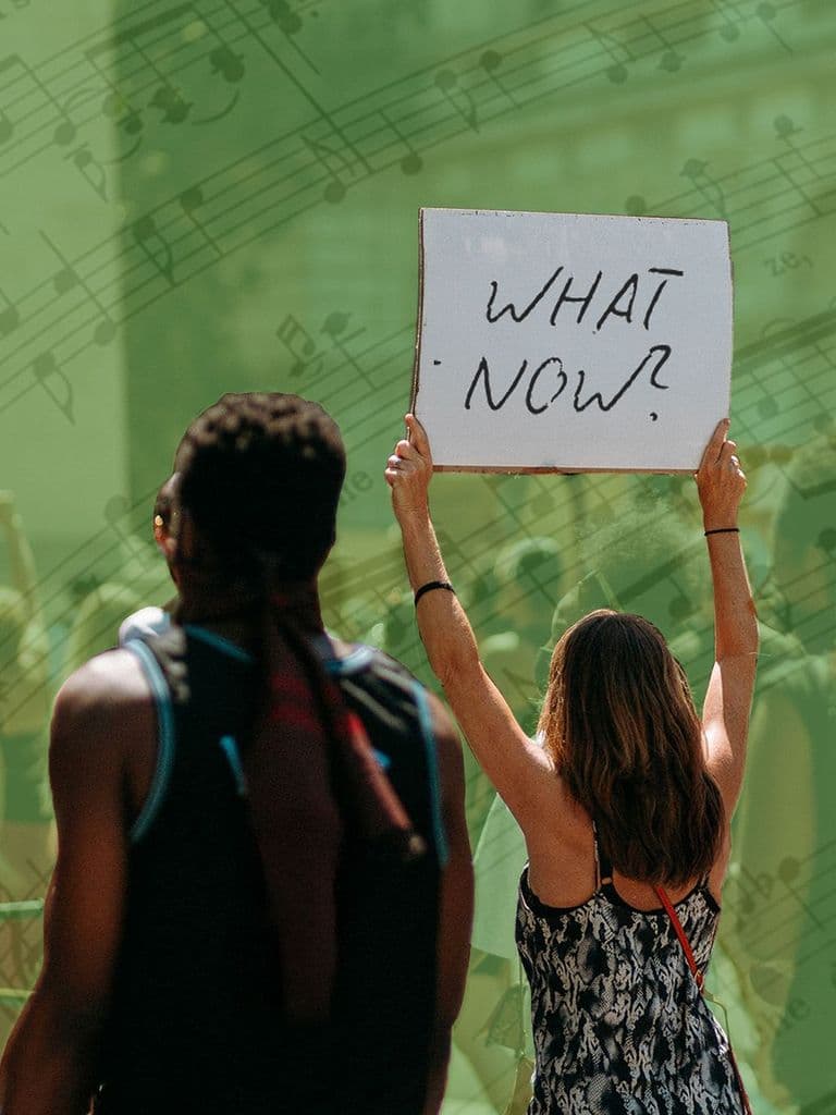 A person holds a sign reading "WHAT NOW?"