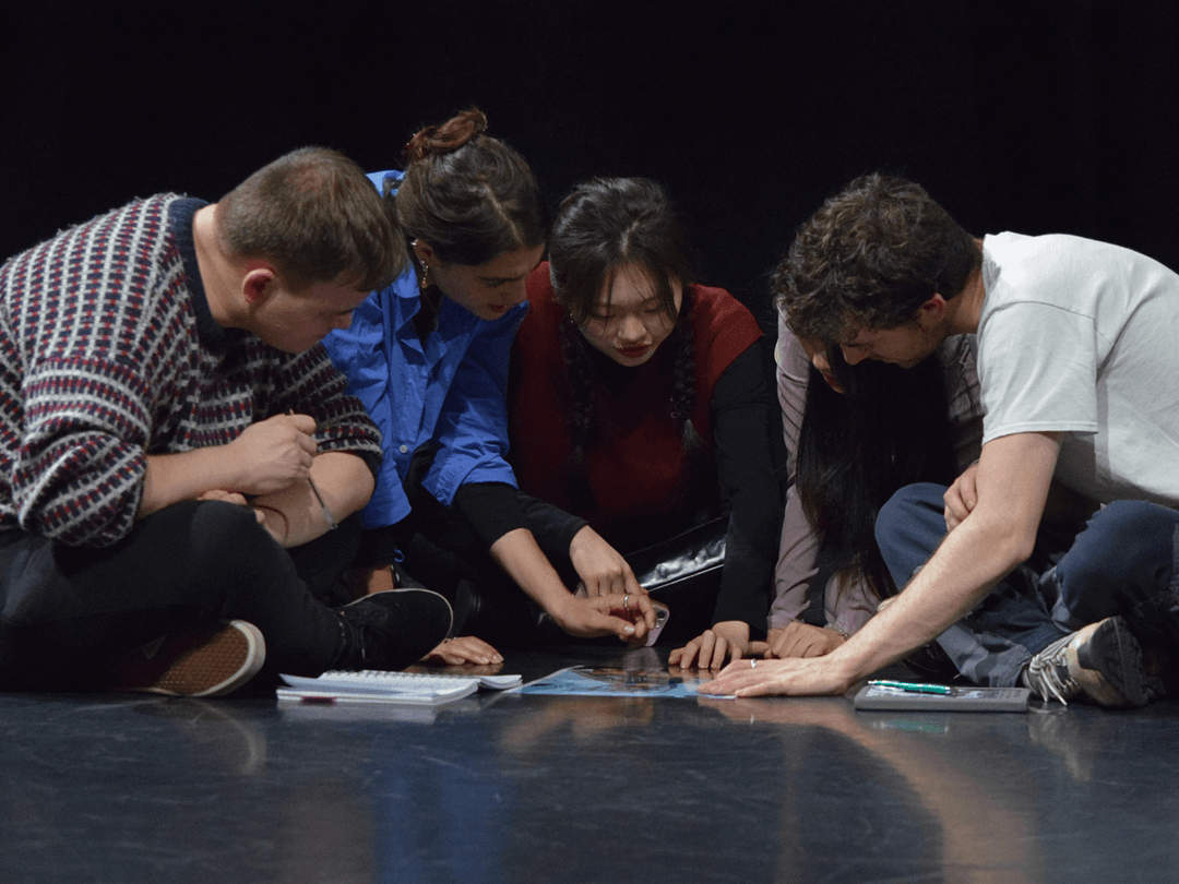 A group of members huddled together on a stage, examining a document spread on the floor with focused expressions.