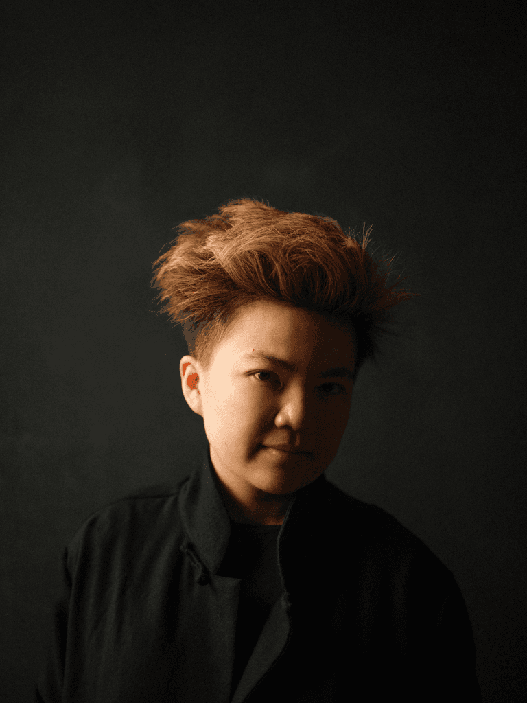 Headshot of Nicola T Chang, who wears a black shirt against a dark background, creating a dramatic silhouette. 