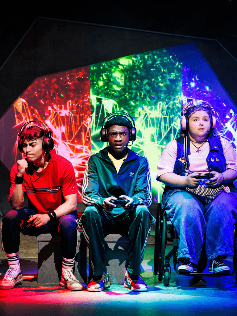 Three young people sit on-stage, facing towards us. Each is backed by a different colour on the set which matches their clothing. They are holding gaming controllers and wearing headsets. 