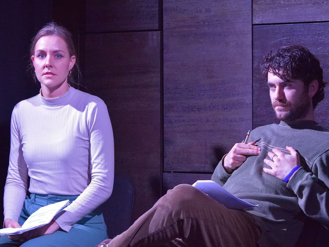 Moana, a woman in a white ribbed top and teal trousers, sits with papers, while Tom, a man in a green jumper holds a pen and appears engaged