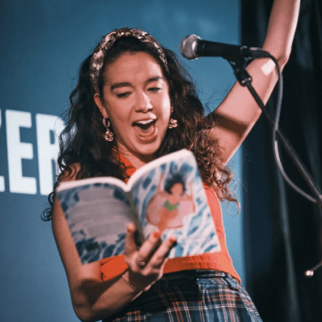 A performer passionately reading from a colorful book at a microphone, in front of a vibrant blue backdrop with text.