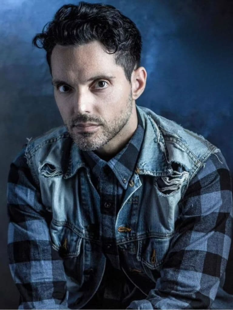 A man with short dark hair and a beard stares intently at the camera. He wears a plaid shirt under a distressed denim jacket, with a moody, blue-toned background.
