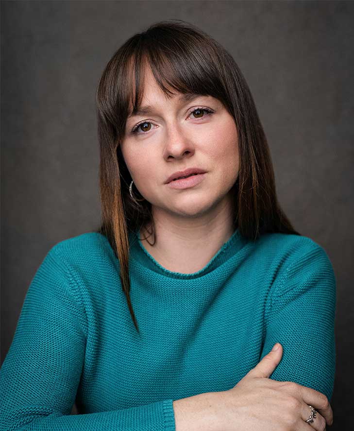 Headshot of Rosie Sheehy – a white woman in a teal shirt with her arms crossed. Her dark brown hair frames her face, with curtain fringe on her forehead.
