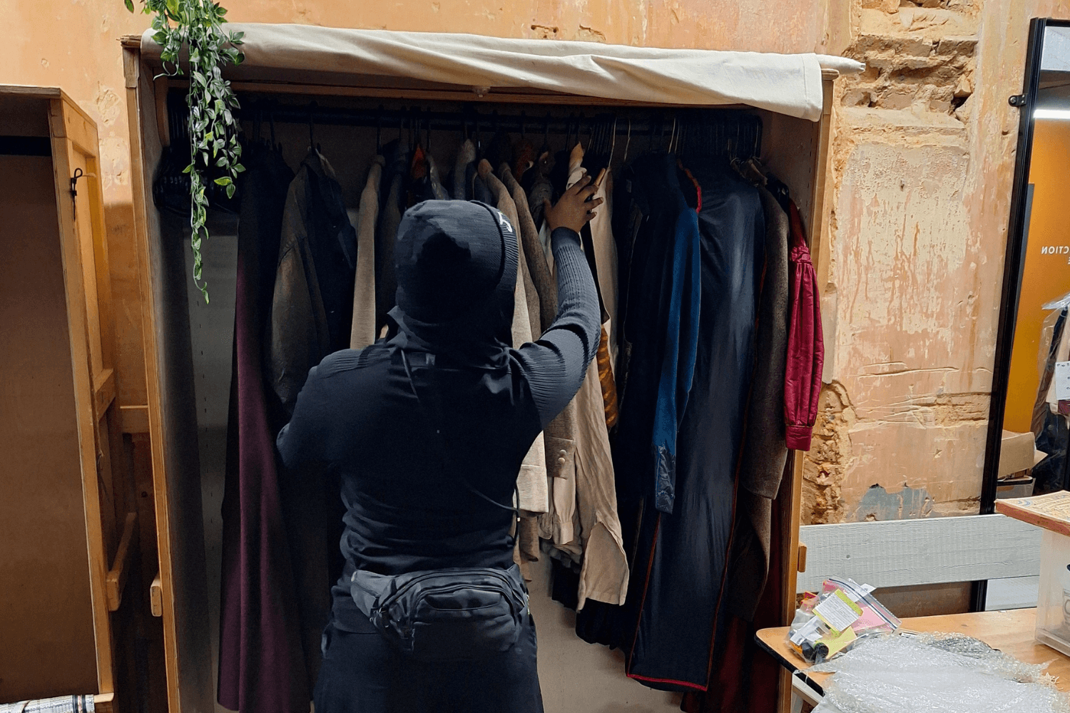 Wardrobe intern, Aisha, a black young woman wearing blacks - looking through the wardrobe at Alexandra Palace.