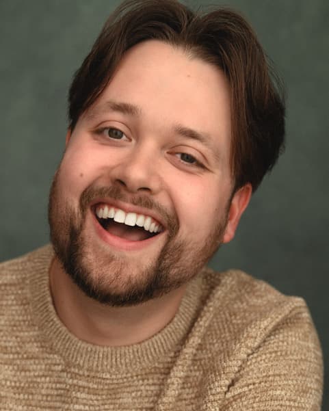 Dan Gill Headshot, a white man with brown hair cut into curtains, a trimmed brown beard and an open smile. Dan is wearing a beige top. 