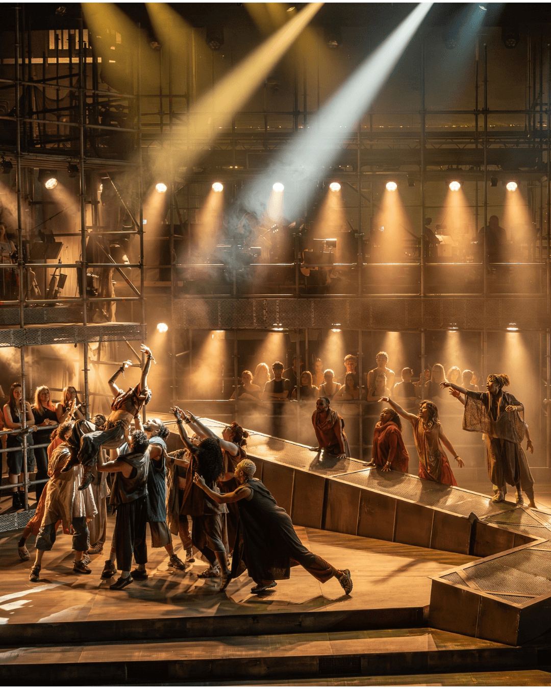 Dramatic theater scene with costumed performers reaching across a stage, bathed in warm amber and white spotlights; energetic, intense mood.
