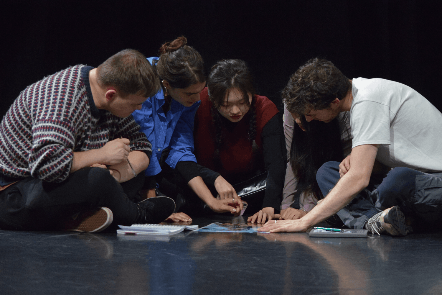 A group of members huddled together on a stage, examining a document spread on the floor with focused expressions.