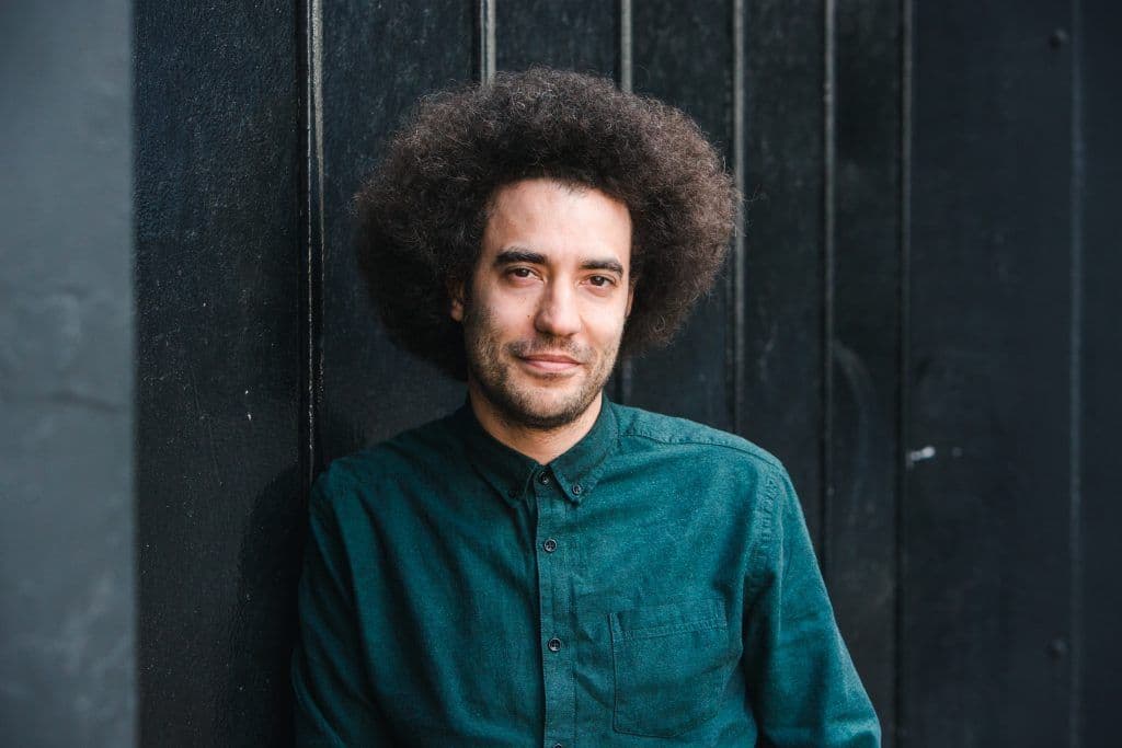 Justin Audibert's headshot, stood in front of black grates with a dark green shirt on.