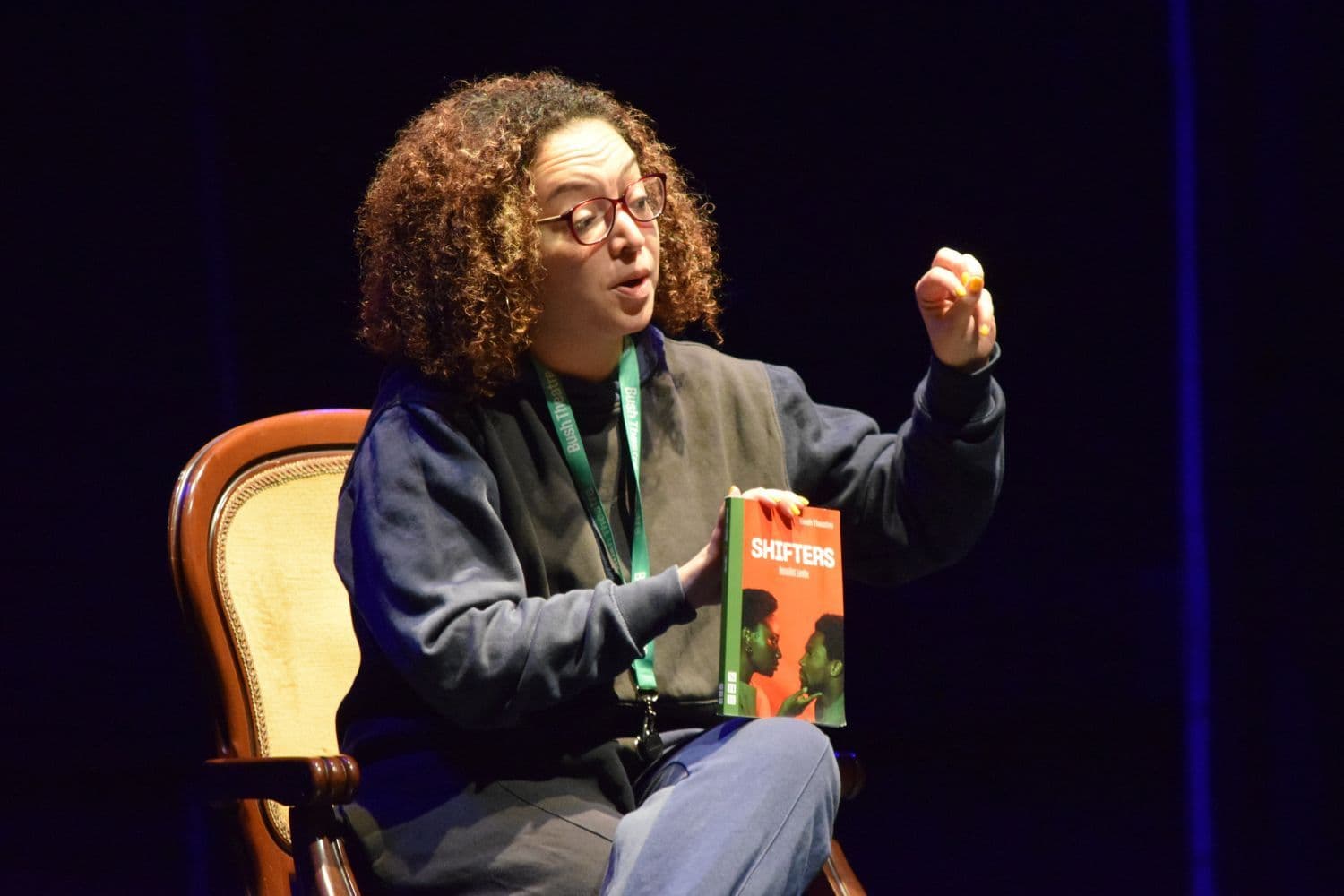 Lynette Linton seated on a chair holding a script titled "Shifters" and gestures while discussing it during a masterclass.