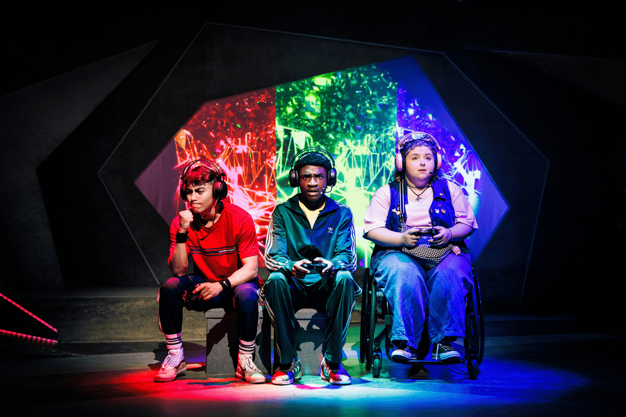 Three young people sit on-stage, facing towards us. Each is backed by a different colour on the set which matches their clothing. They are holding gaming controllers and wearing headsets. 