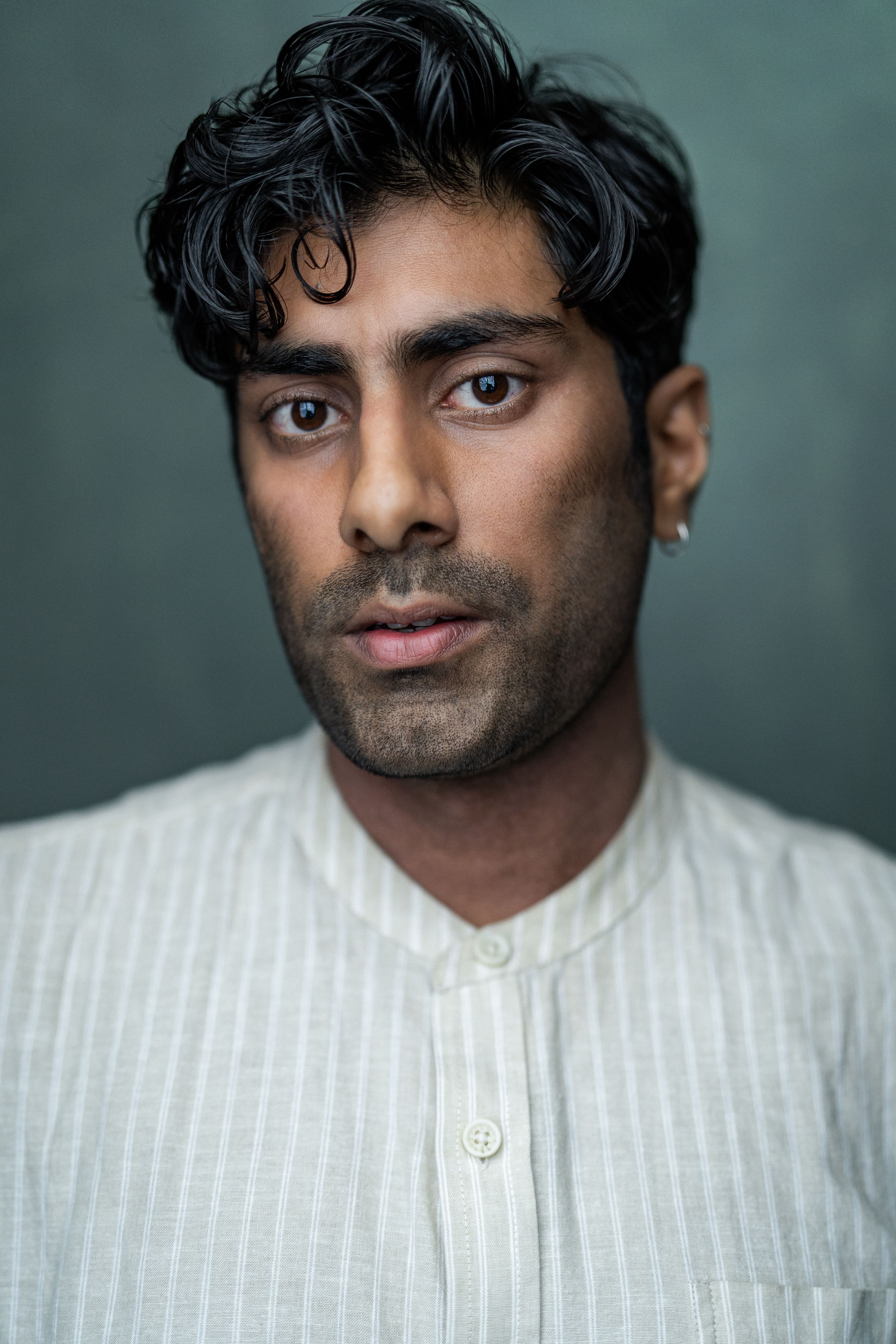 Rishi Rian headshot. An asain man with curly black hair and styled stubble. Wearing a white striped shirt.