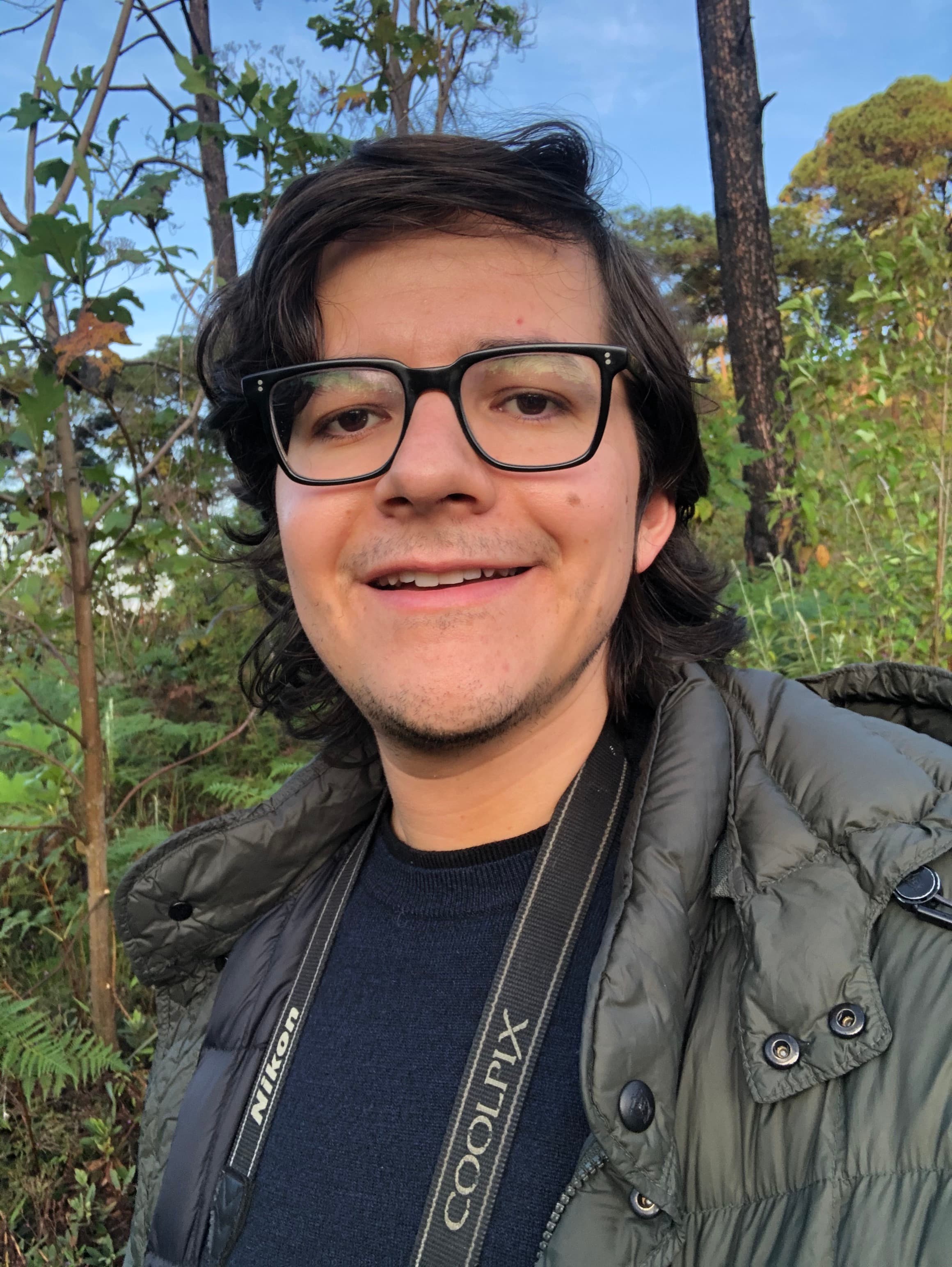 Santiago Guerra Headshot, infront of forest background. A man with brown hair and black rimmed glasses and an open smile.