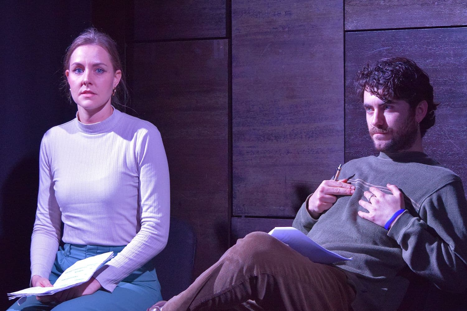 Moana, a woman in a white ribbed top and teal trousers, sits with papers, while Tom, a man in a green jumper holds a pen and appears engaged