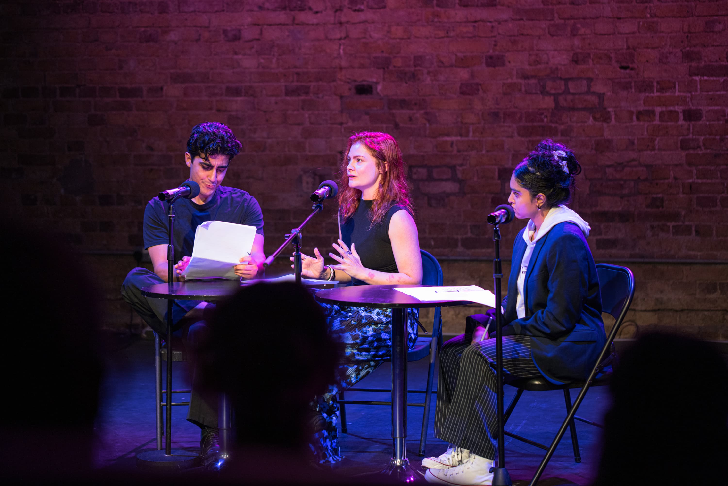 Three young people sit at a table with microphones and scripts in front of them. They are acting out a scene.