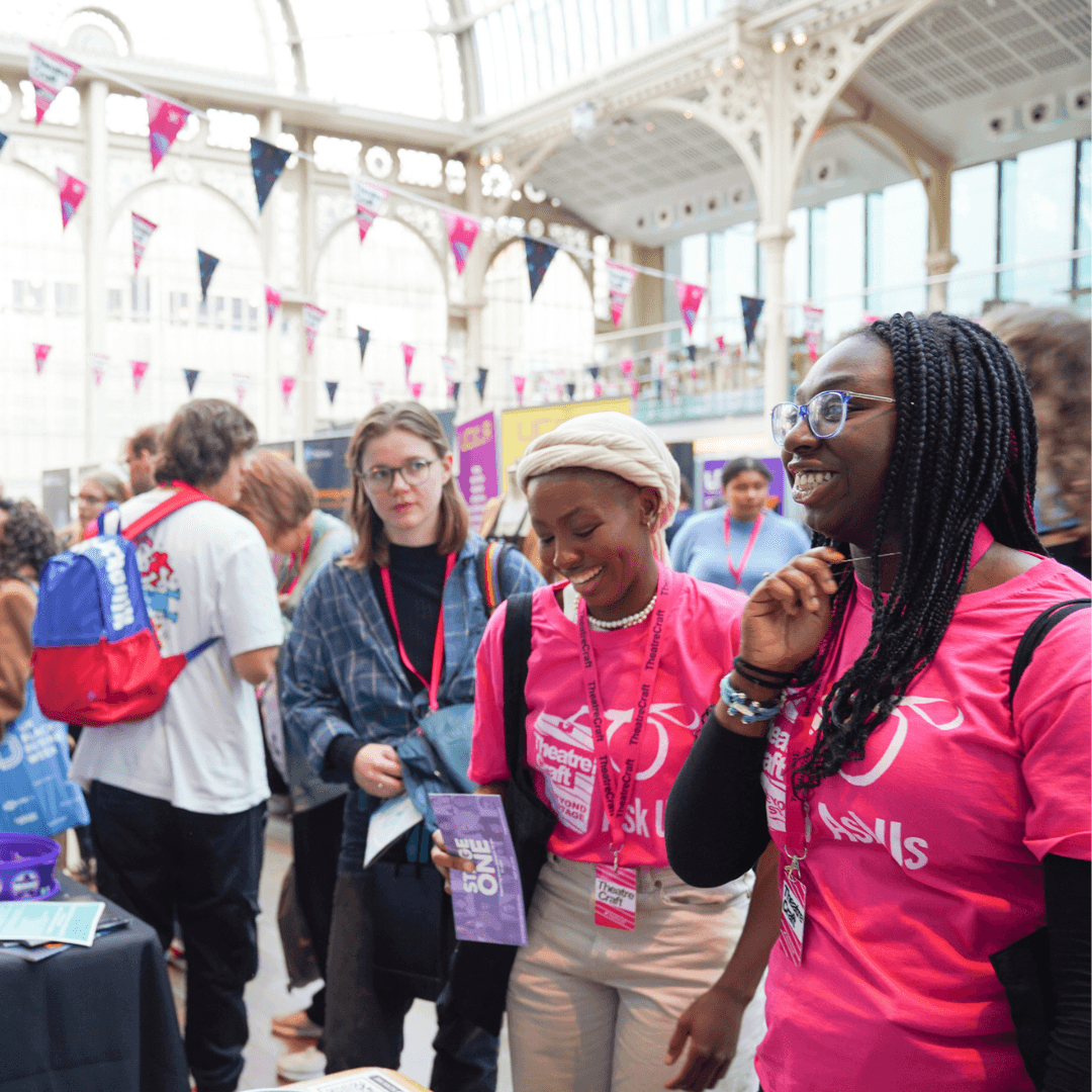Two TheatreCraft volunteers in pink t-shirts, with bunting overhead and engaged in conversation.