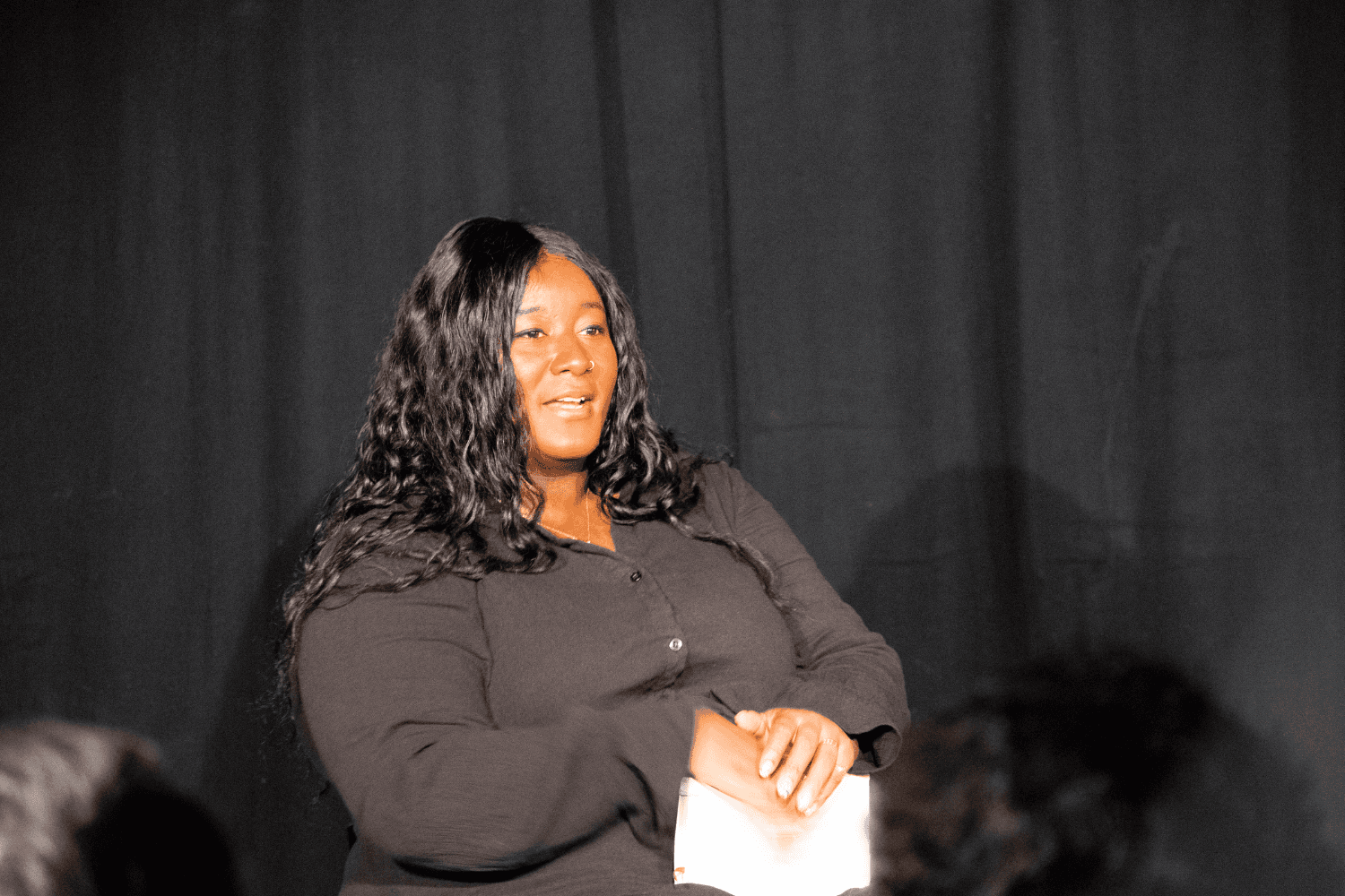 Yolanda Mercy sat on a chair with a notebook in her hands. A black woman with long black wavey hair, wearing a black buttoned dress.