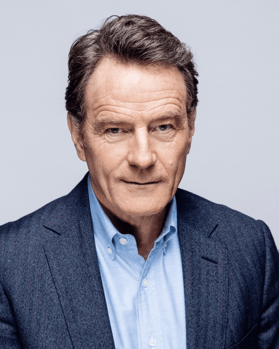 Headshot of Bryan Cranston, a man with neatly styled hair wearing a tailored dark blazer over a light blue shirt, set against a plain light grey background.
