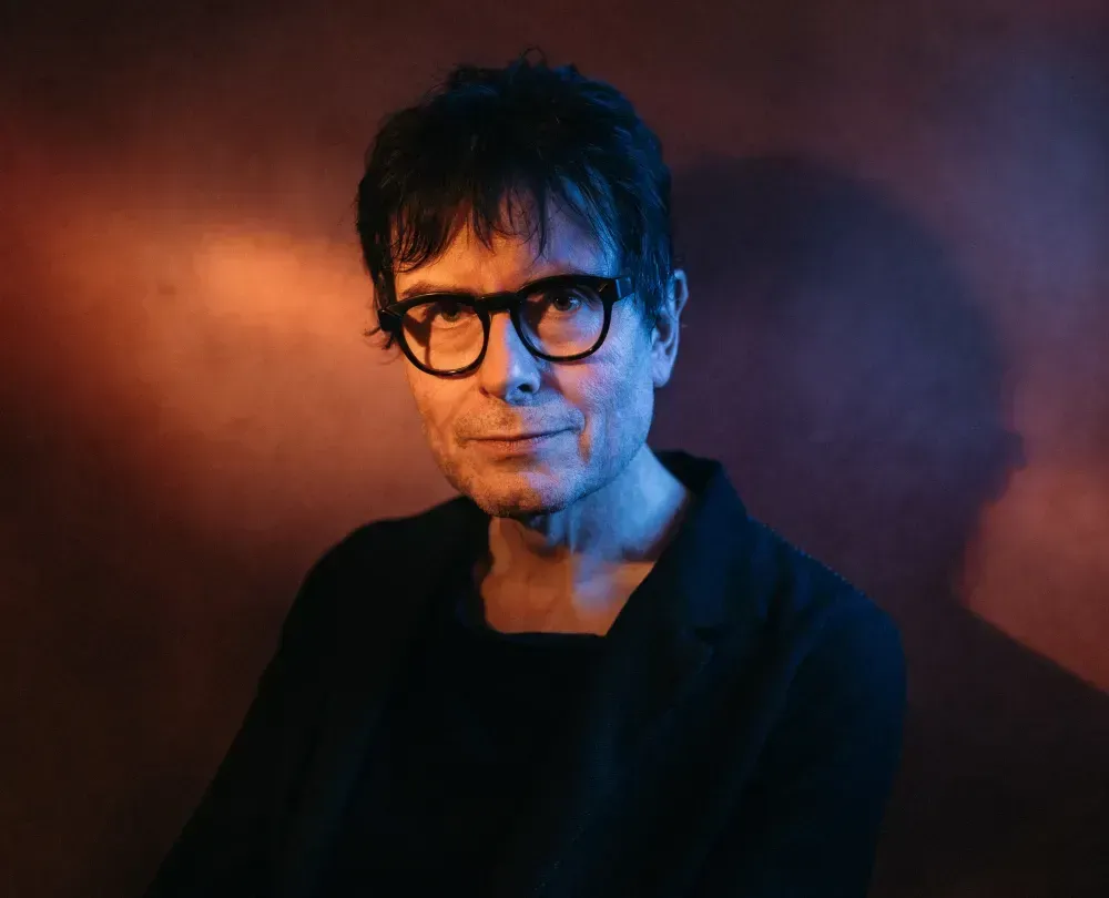 Director, James Macdonald in black rimmed glasses and dark clothes, standing against a background which is lit to shimmer gold and purple. Light shining on to James' face giving a soft glow.