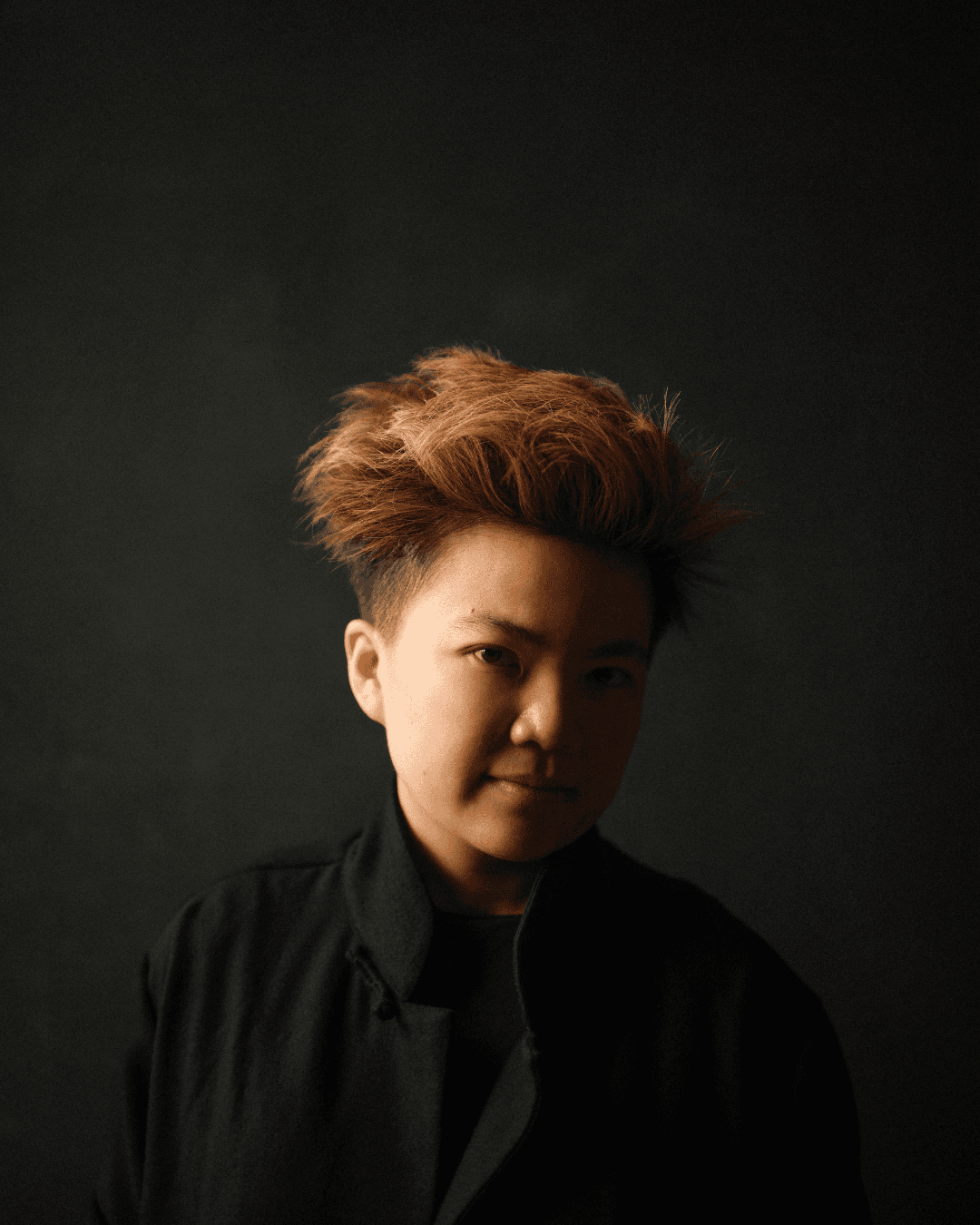 Headshot of Nicola T Chang, who wears a black shirt against a dark background, creating a dramatic silhouette.