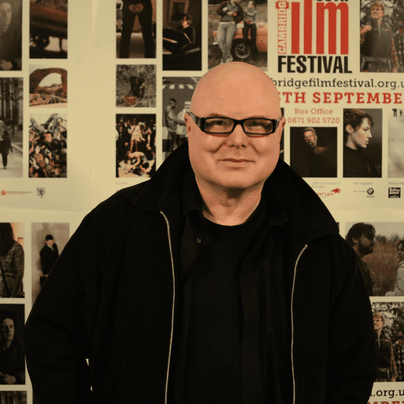 Philip Ridley dressed in black, wearing black rimmed glasses in front of a selection of film posters.