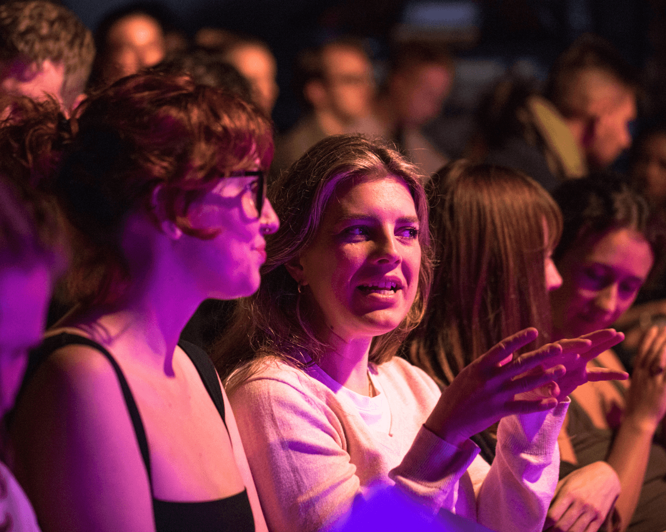 A group of people sit closely together at a theatre, with one woman in the center gesturing as she speaks to the person next to her.