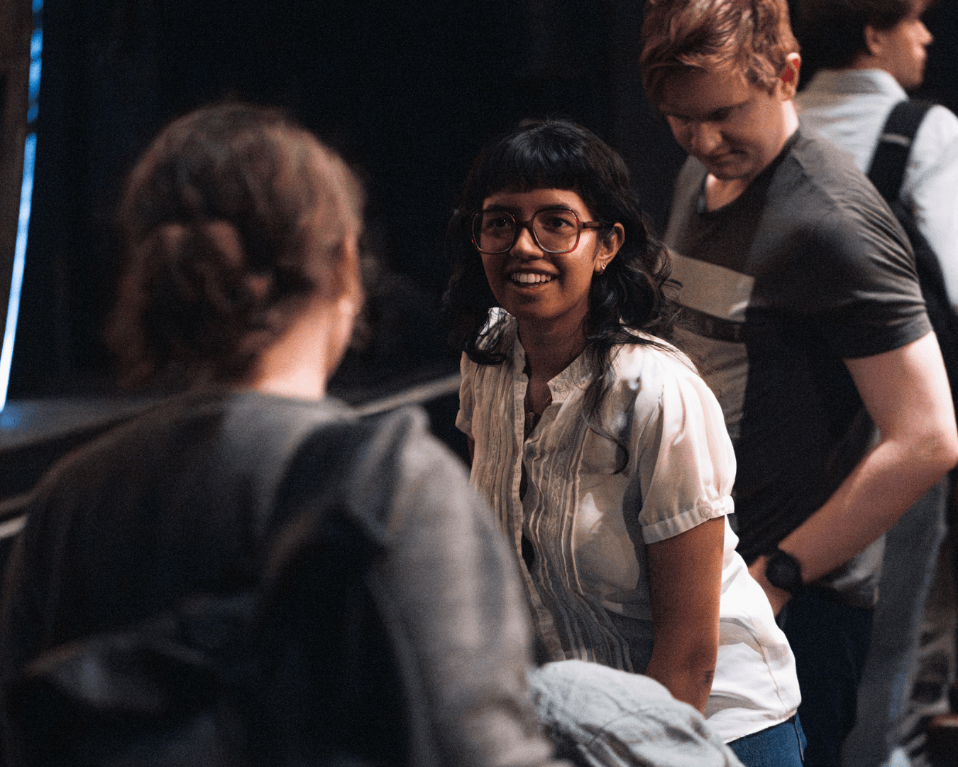 A woman with glasses and a white shirt smiles while talking to another person. A person in a striped shirt stands infront of her. The setting is in a theatre.