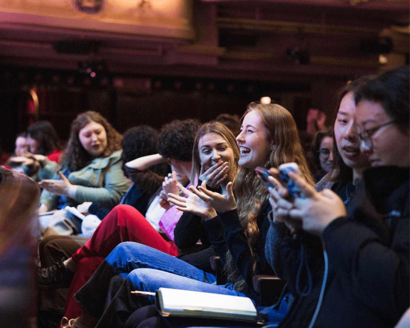 Audience members in theatre seats engage by laughing, clapping and using phones during an event in a dimly lit venue.