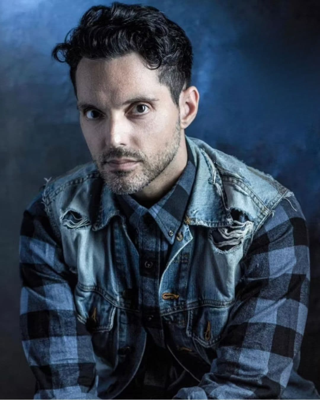 A man with short dark hair and a beard stares intently at the camera. He wears a plaid shirt under a distressed denim jacket, with a moody, blue-toned background.