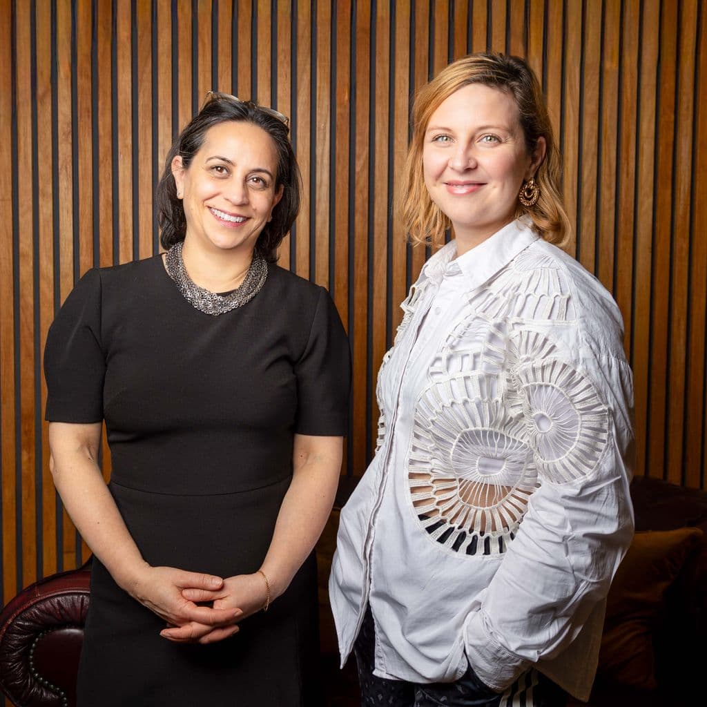Clare Annamalai and Alex Duarte-Davies, two female-identifying and presenting individuals standing indoors against a wooden slat wall; Clare is in a black dress, Alex is in a white shirt with lace details.