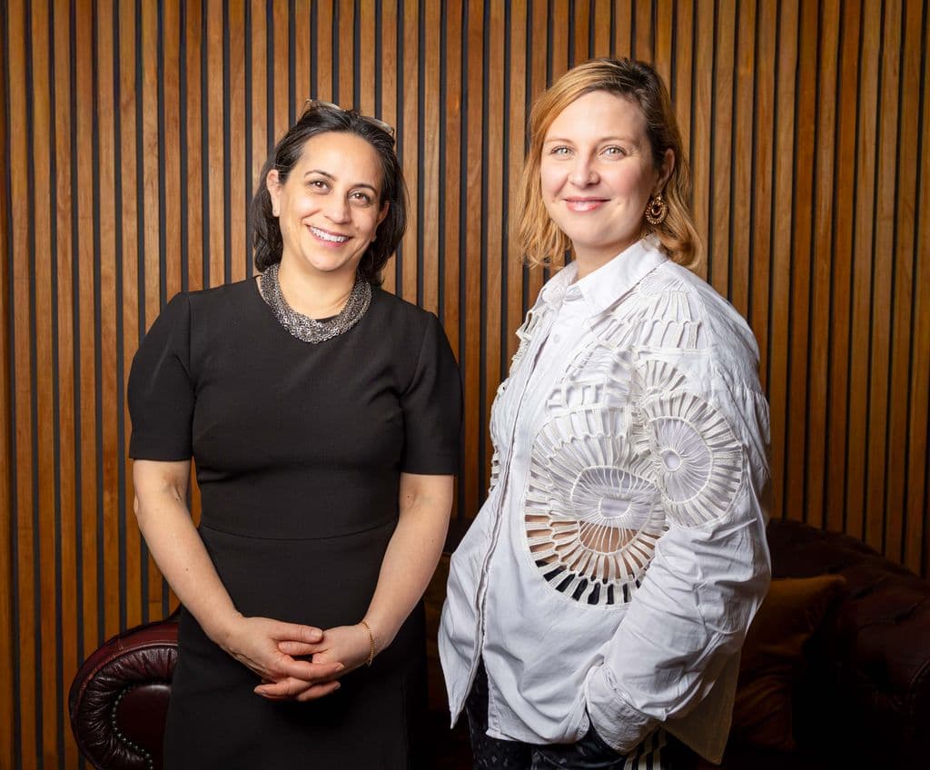 Clare Annamalai and Alex Duarte-Davies, two female-identifying and presenting individuals standing indoors against a wooden slat wall; Clare is in a black dress, Alex is in a white shirt with lace details.