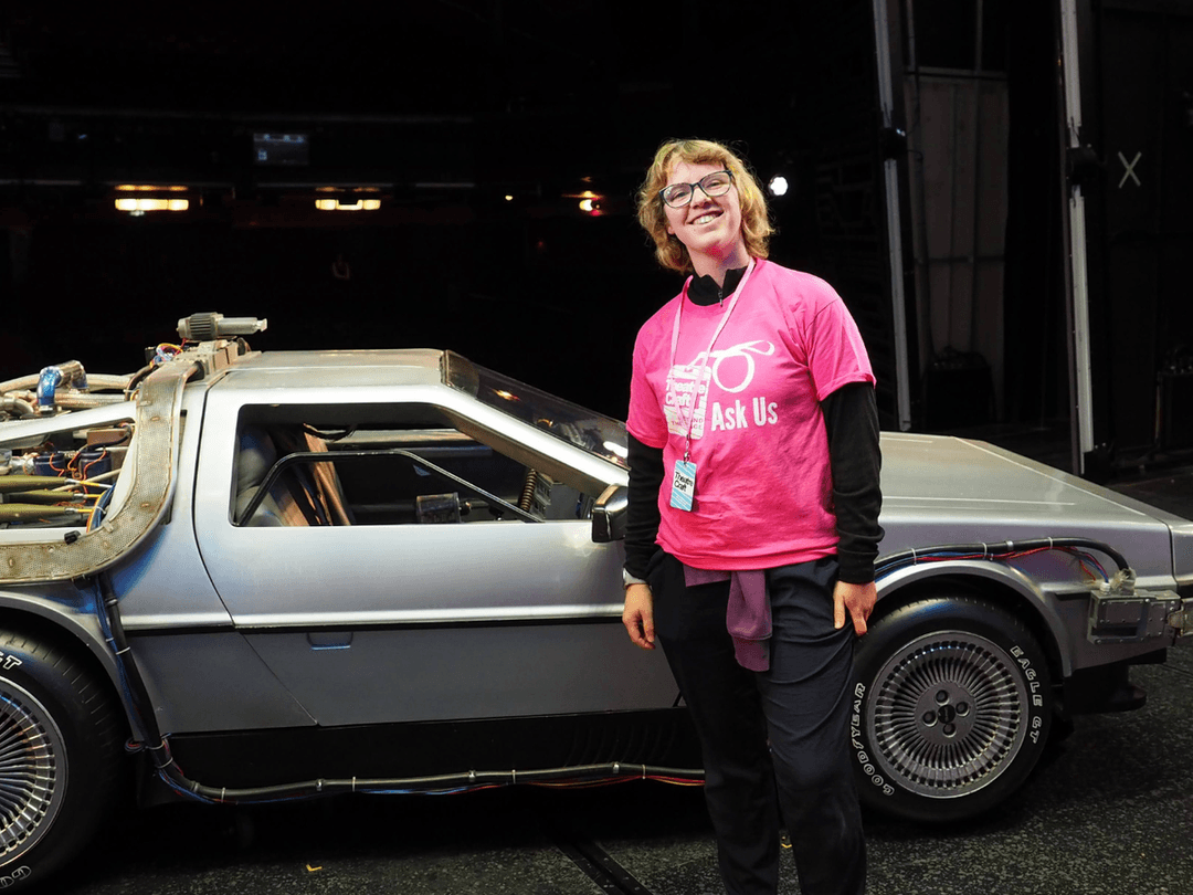 Imogen at Back To The Future TheatreCraft. A white woman with short blonde hair, wearing a TheatreCraft volunteer T-Shirt in front of the infamous car.