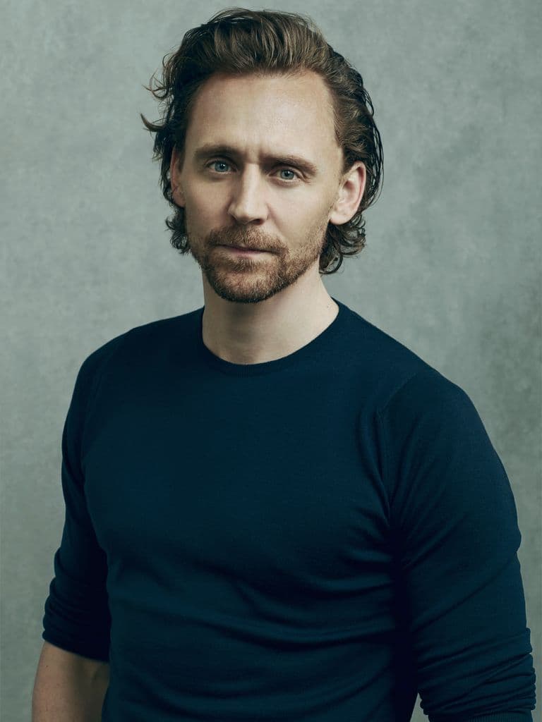 Tom Hiddleston, White male actor with brown hair and trimmed beard. Wearing a dark blue long sleeved T-shirt against a green background.