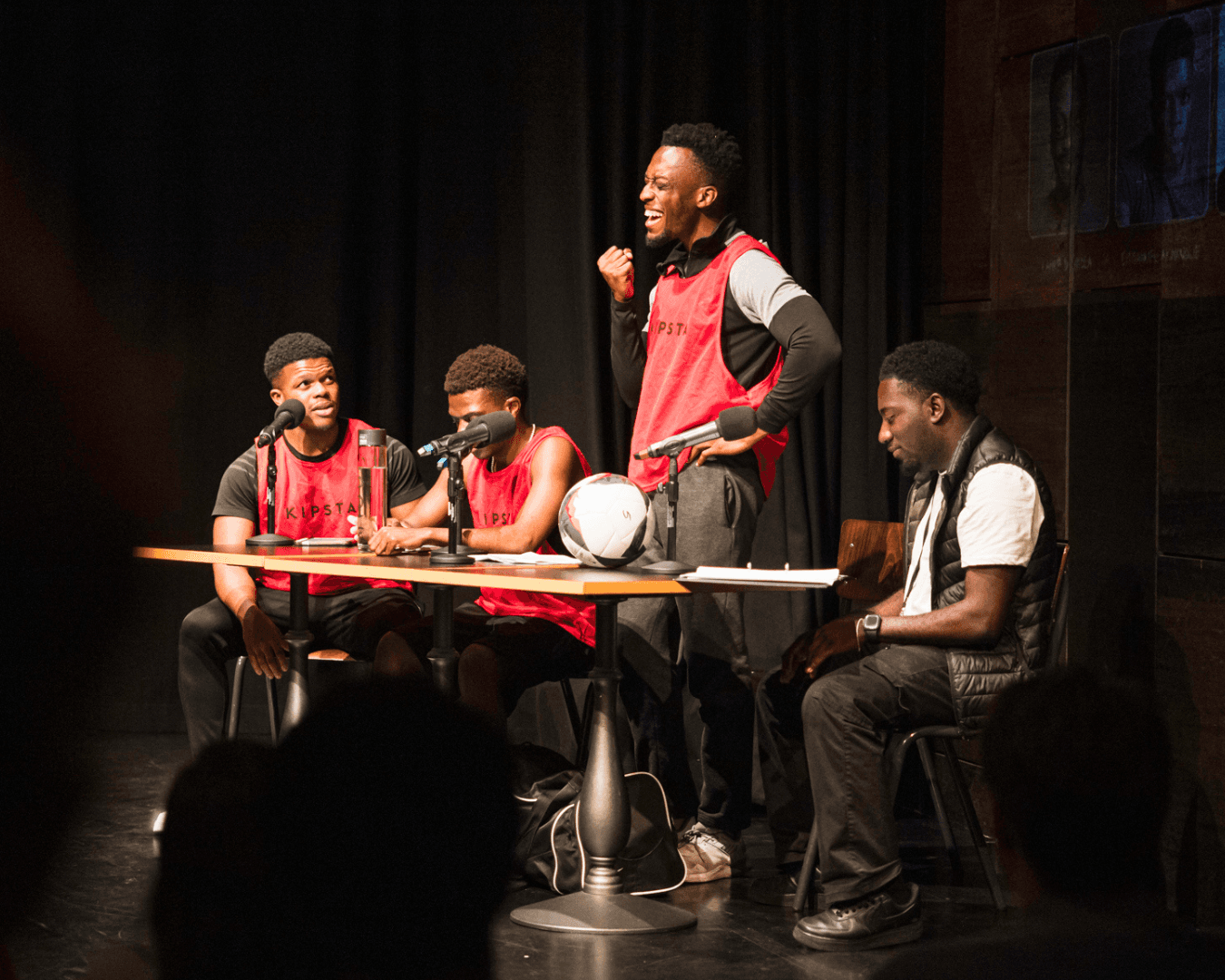 Four men on stage, three wearing red sports vests, engaged in discussion around a table with microphones and a football.