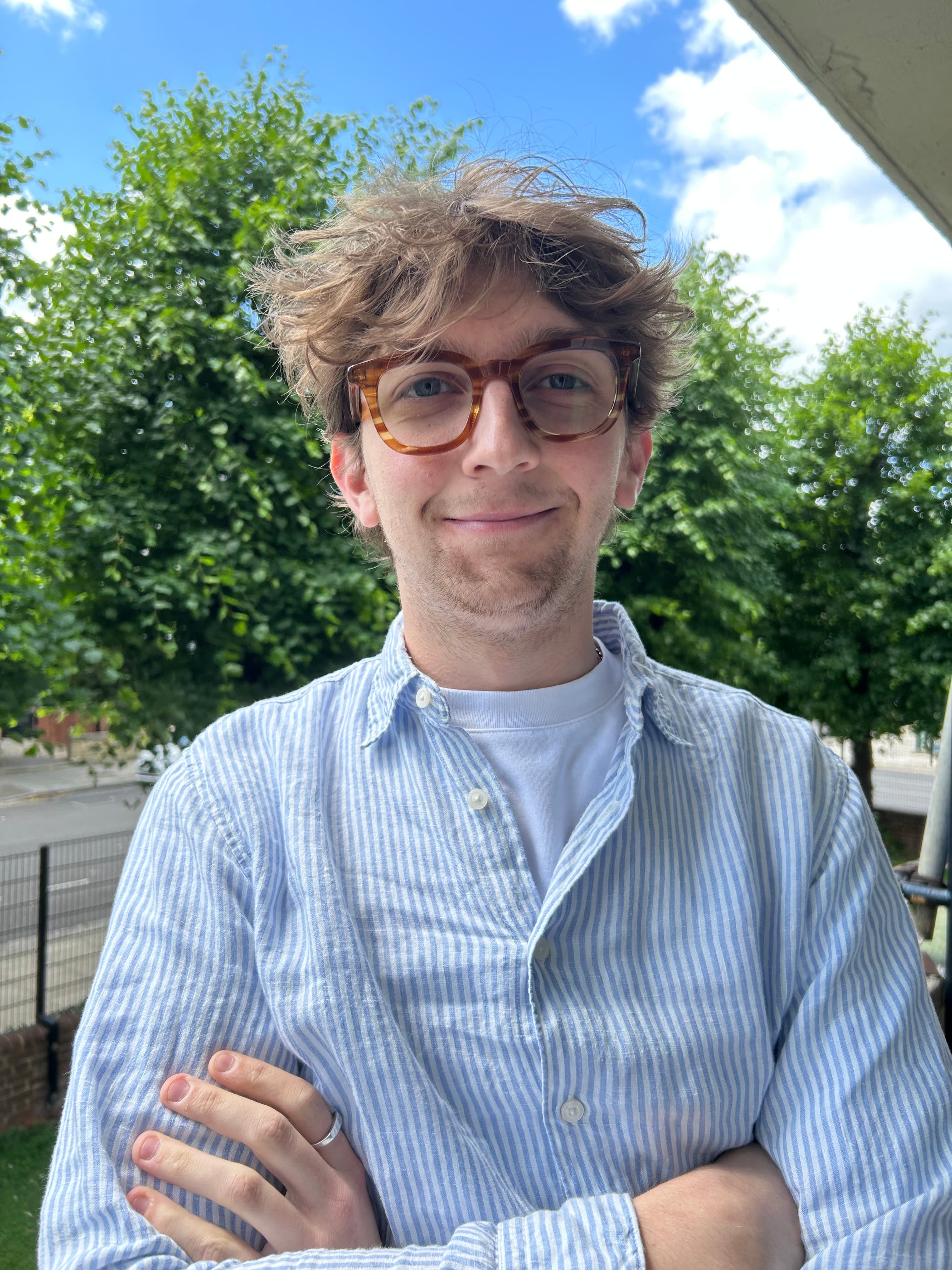 Charles, a white male with tousled hair wearing a blue and white striped shirt, stands outdoors with arms crossed, surrounded by green trees and blue skies.