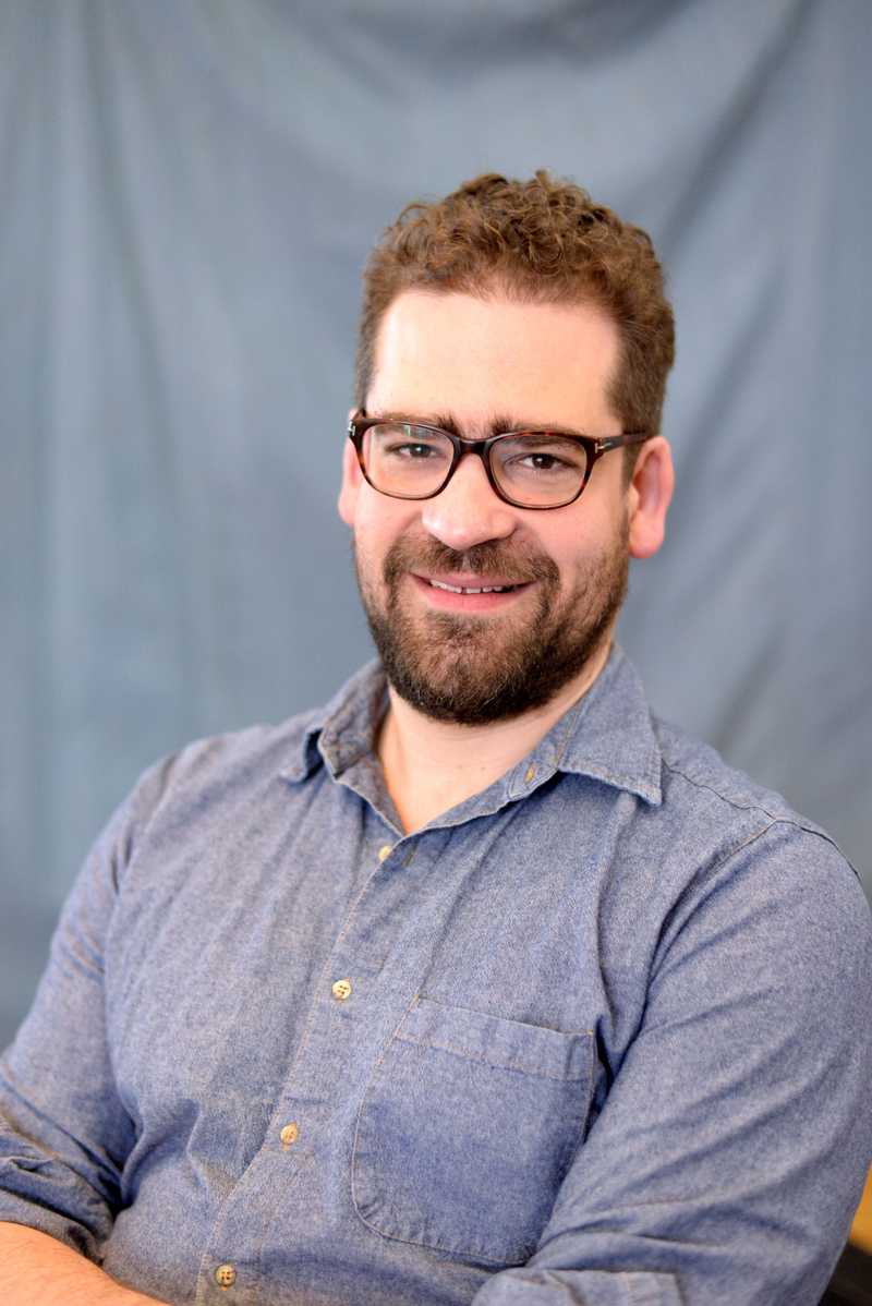 Headshot of Max Webster taken against a material grey/blue background. Max is wearing a blue shirt with a collar. He is wearing a brown pair of glasses and has brown hair and facial hair.
